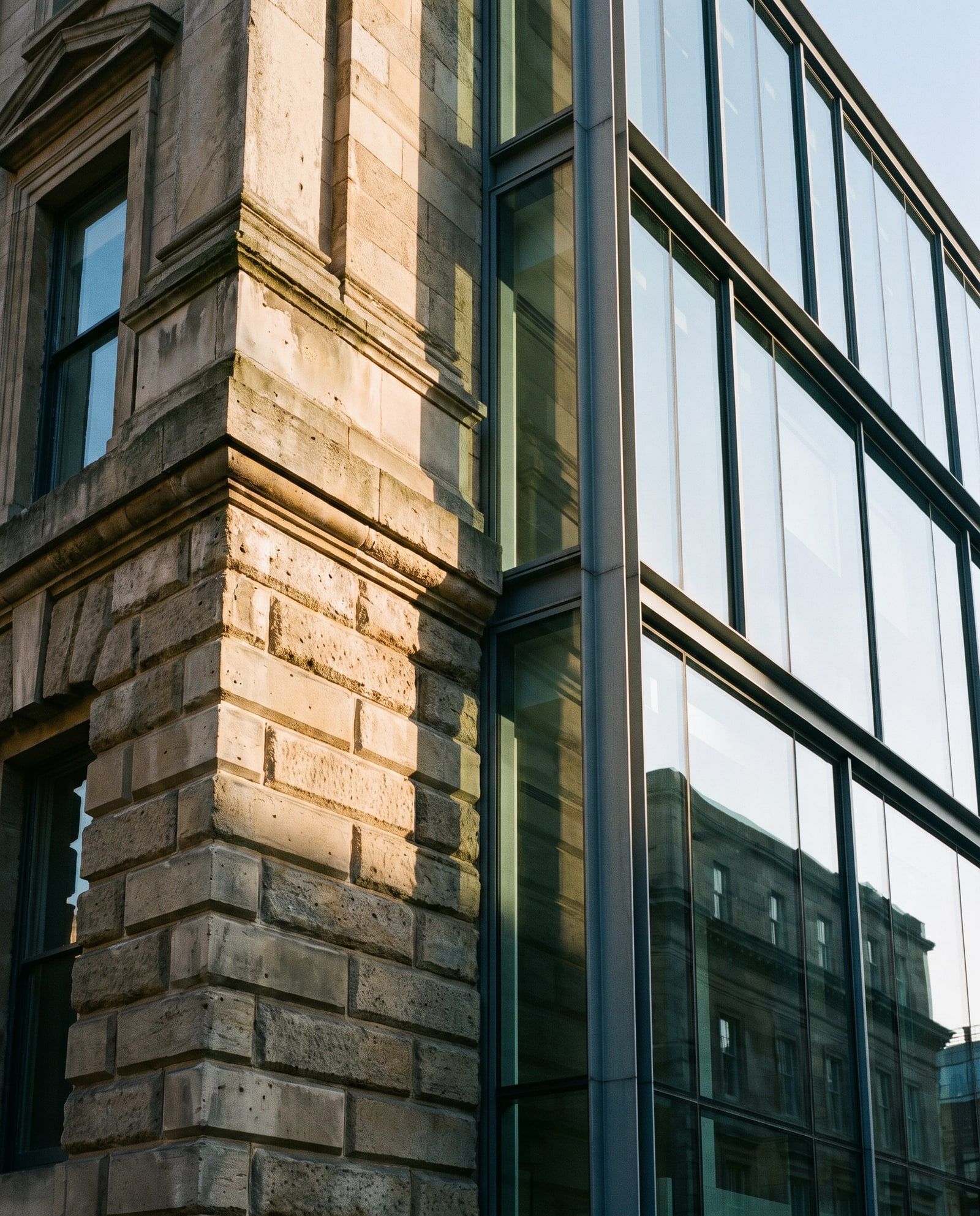 The corner of a historic stone facade meeting a new glass-and-steel extension in low sunlight.